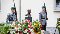 Zwei Soldaten stehen mit Gewehr neben einer Büste und hinter Blumenkränzen.