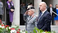 Ein Soldat und ein Mann im Anzug stehen vor mehreren Blumenkränzen. Der Soldat salutiert.