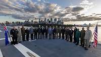 Menschen in Uniform stehen im Halbkreis auf einem Schiff. Im Hintergrund ist die Skyline von Sydney.