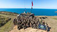 Gruppenbild mit zahlreichen Soldaten und einem Fahrzeug, im Hintergrund das Meer..