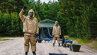 Mehrere Soldaten in einem ABC-Schutzanzug stehen hintereinander auf einem Schotterweg im Wald. 