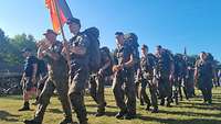 Soldaten marschieren mit Gepäck und der deutschen Truppenfahne über eine Wiese