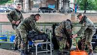 Vier Soldaten stehen an der Wasserkante von einem Gewässer. Um sie herum liegen Boxen und Kabel.