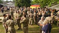 Zahlreiche Soldaten und Zivilisten stehen zwischen Bänken und Tischen auf einer Wiese.