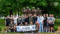 Eine Gruppe von Zivilisten und Soldaten steht mit einem Banner vor einer Statur