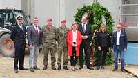 Vor einem großen Rohbau stehen Frauen und Männer in Zivil und Uniform an einem Richtkranz