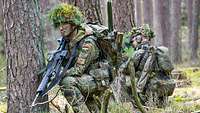 Zwei Soldaten hocken getarnt und bewaffnet im Wald hinter Baumstämmen.