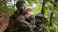 Ein Zivilist in Flecktarnparka schaut durch ein Messgerät. Ein Soldat steht daneben. 