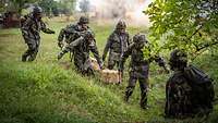 Vier Soldaten schleppen Munitionskisten aus Holz über eine Wiese.