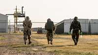 Drei Soldaten in Schutzanzügen gehen auf entfernte Gebäude zu.