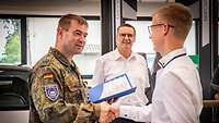 Ein Soldat und ein Jugendlicher in hellem Hemd stehen sich gegenüber und geben sich die Hand.