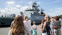 Viele Menschen stehen vor einem grauen Schiff, von dem Personen in sandfarbenen Uniform winken.
