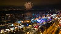 Seegelschiffe in einem Hafen bei Nacht aus der Vogelperspektive. Im Hintergrund ein Feuerwerk. 