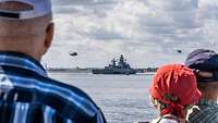 Ein Schiff fährt auf einem Gewässer. Über dem Schiff fliegen zwei Hubschrauber. 