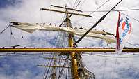 Ein Mast eines Segelschiffes ist zu sehen. Darauf befinden sich Personen, die das Segel einholen.