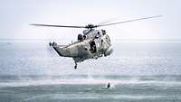 Ein Hubschrauber fliegt über ein Gewässer. Aus dem Hubschrauber springt ein Mensch in das Wasser.