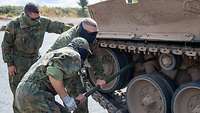 Zwei Soldaten schrauben gemeinsam an einer Panzerkette. Ein weiterer Soldat steht daneben