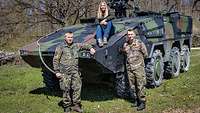 Eine Frau sitzt auf einem Panzer. Zwei Männer in Uniform stehen davor.