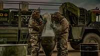 Two people wearing protective suits and masks are emptying a plastic bag over a tapered vat.