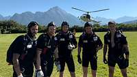 Fünf Personen stehen in Fallschirmspringeroutfits auf einer Wiese