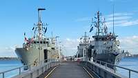 Im Vordergrund ein massiver Anleger, links und rechts davon zwei Kriegsschiffe der Marine.