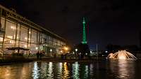 Bei Nacht: Ein Gebäude mit Glasfront, ein Wasserbecken mit Fontäne, der grün illuminierte Funkturm 