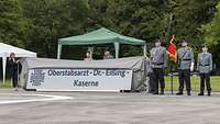 Vier Personen enthüllen das neue Kasernenschild in Kronshagen. Rechts daneben steht eine Ehrenabordnung mit Truppenfahne