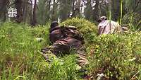 Zwei Soldaten kriechen durch Moos und hohes Gras in einem Wald.