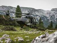 Ein NH-90 schwebt über eine Wiese zwischen den Alpen, dabei schaut ein Soldat aus der Seitentür.