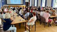 Mehrere Personen sitzen in einem großen Raum an Tisch und schauen auf eine Leinwand. 