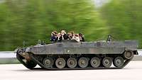 Aus einem fahrenden Panzer ohne Panzerturm schauen mehrere junge Frauen heraus.