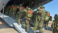 Mehrere Soldaten betreten in Zweierreihe mit Fallschirmen im Gepäck auf dem Rücken ein Flugzeug.