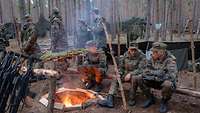Mehrere Soldaten sitzen um ein Feuer herum, im Hintergrund aufgebaute Zeltplanen.
