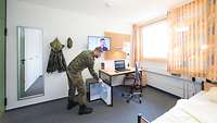 Blick in eine Stube mit Bett, Schreibtisch, Fernseher und einem Kühlschrank, den ein Soldat öffnet.
