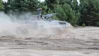 Ein Panzer fährt von links nach rechts durch tiefen Sand vor einem Wald