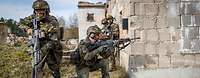 Drei bewaffnete Soldaten stehen und hocken nahe einer Hausmauer und sichern nach allen Seiten.