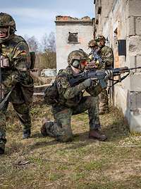 Drei bewaffnete Soldaten stehen und hocken nahe einer Hausmauer und sichern nach allen Seiten.