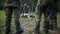 Eine Person liegt mit entblößtem Oberkörper auf dem Boden, ein Soldat hilft ihm beim Entkleiden