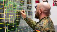 Soldat mit Glatze sitzt vor einem Blackboard und füllt mit grünem Stift eine Fluginformationstafel aus.
