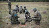 Mehrere Soldaten knien auf einem Boden vor einer Sprengmine, die rot umrandet ist