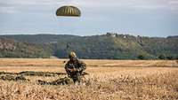 Ein Fallschirmjäger hockt nach der Landung auf einem geernteten Feld und schaltet das Funkgerät ein.