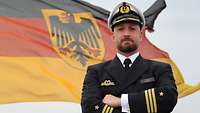 Marineoffizier mit dunkelblauer Uniformjacke und Schirmmütze vor einer deutscher Flagge. Die Arme vor der Brust verschränkt.