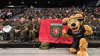 Vielen Soldaten sitzen im Publikum auf einer Tribüne mit einer Flagge. Ein Maskottchen kniet davor. 