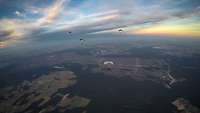 Mehrere Fallschirmjäger fliegen in der Dämmerung mit dem letzten Licht am Himmel.
