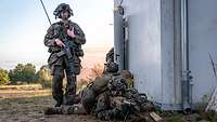 Soldaten liegen an einer Hauswand mit ihren Waffen am Boden, ein Soldat steht bewaffnet daneben.