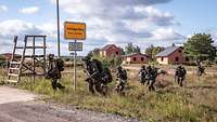 Eine Gruppe von Soldaten überquert mit schnellen Schritten eine Straße vor einem Ortseingangsschild.