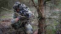 Ein Soldat hockt mit einer Waffe in der Hand hinter einem Baum.