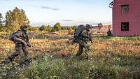 Soldaten laufen mit der Waffe in der Hand schnell über eine Wiese an einem Haus vorbei.