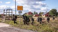 Soldaten mit Gepäck und Waffe stürmen über eine Straße, im Hintergrund Häuser.