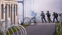 Soldaten stürmen über eine Straße. Stacheldraht ist auf der Straße ausgerollt.